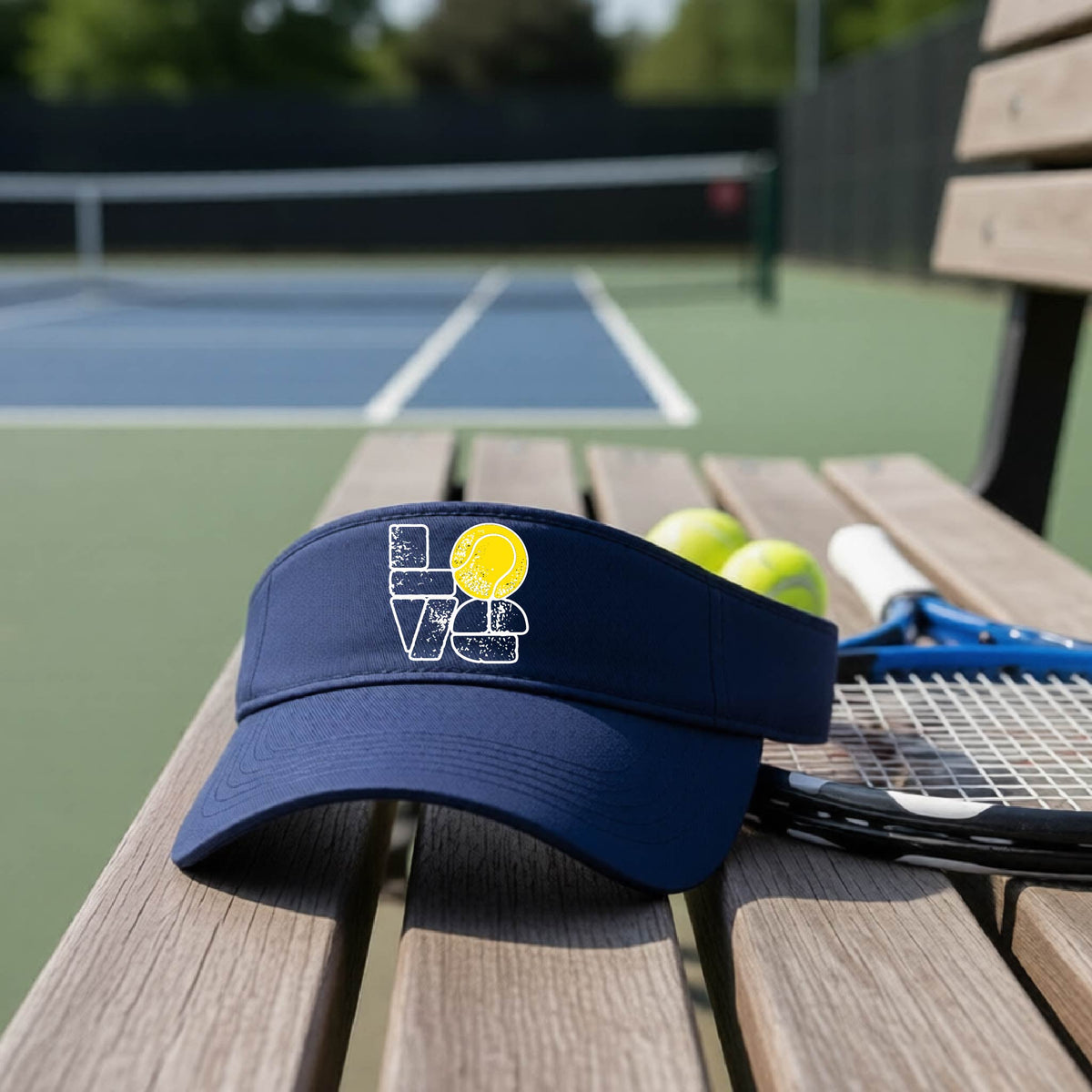 A navy blue visor with a white and yellow tennis-themed graphic "LOVE" with a tennis ball replacing the O, sits on a wooden bench at a tennis court, with tennis balls and a racket in the background.