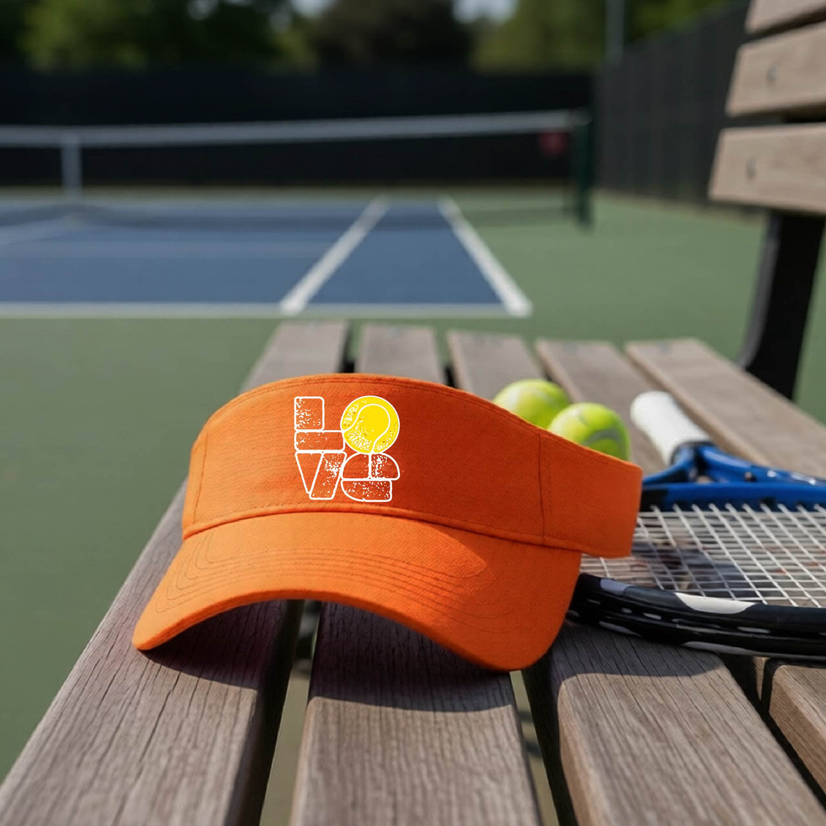 An orange visor with a white and yellow tennis-themed graphic "LOVE" with a tennis ball replacing the O, sits on a wooden bench at a tennis court, with tennis balls and a racket in the background.