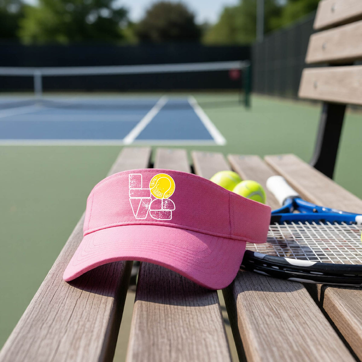 A pink visor with a white and yellow tennis-themed graphic "LOVE" with a tennis ball replacing the O, sits on a wooden bench at a tennis court, with tennis balls and a racket in the background.