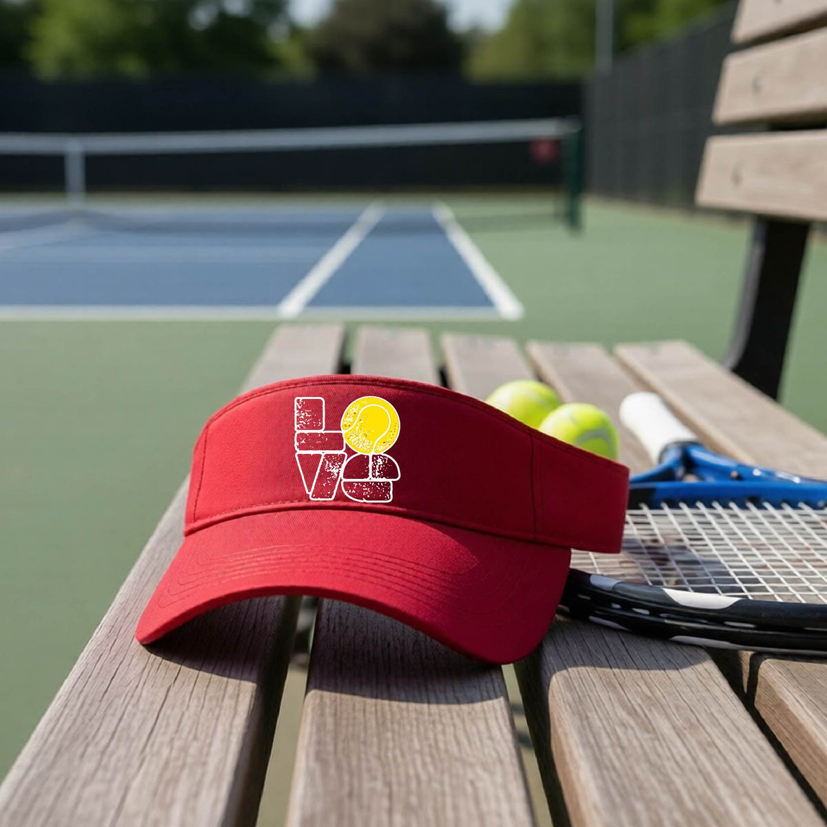 A red visor with a white and yellow tennis-themed graphic "LOVE" with a tennis ball replacing the O, sits on a wooden bench at a tennis court, with tennis balls and a racket in the background.