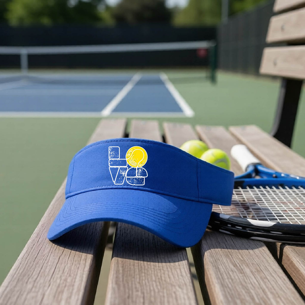 A royal blue visor with a white and yellow tennis-themed graphic "LOVE" with a tennis ball replacing the O, sits on a wooden bench at a tennis court, with tennis balls and a racket in the background.