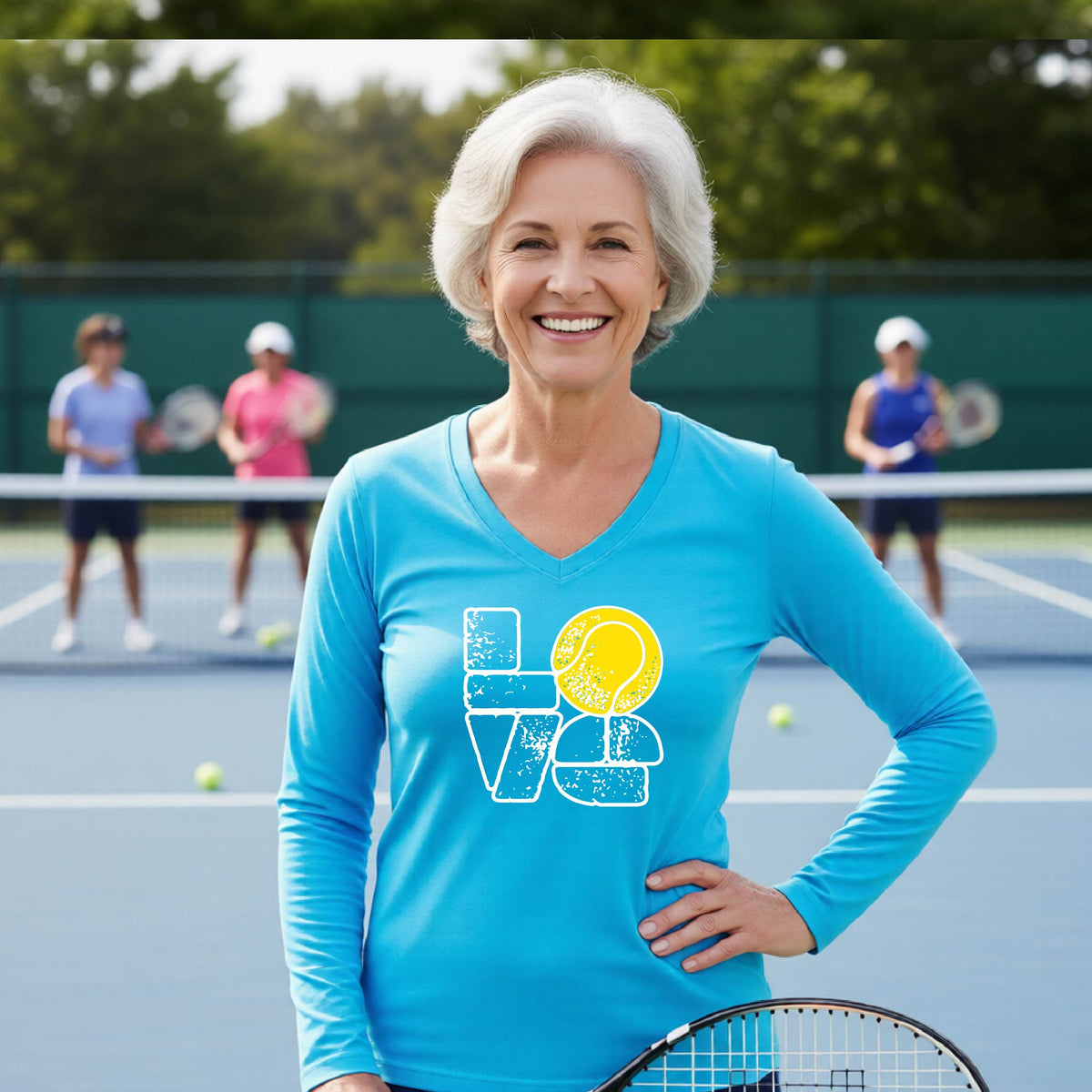 A woman wearing a bright blue long-sleeve t-shirt with a white and yellow tennis-themed graphic "LOVE" with a tennis ball replacing the O, with a tennis  court and players in the background.