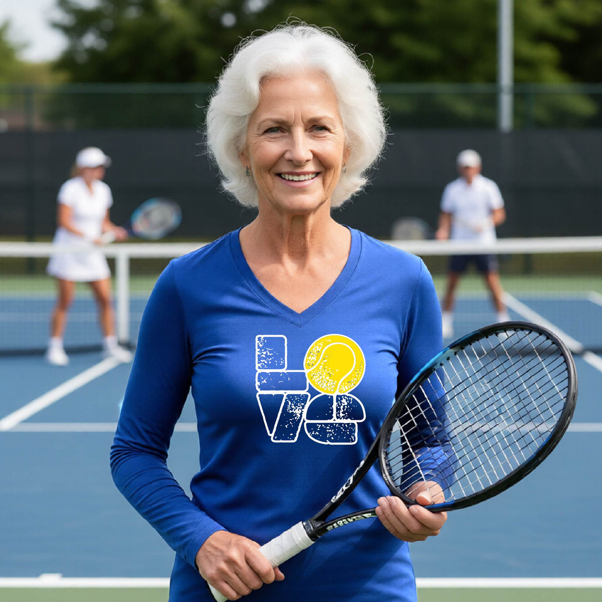 A woman wearing a royal blue long-sleeve t-shirt with a white and yellow tennis-themed graphic "LOVE" with a tennis ball replacing the O, with a tennis  court and players in the background.