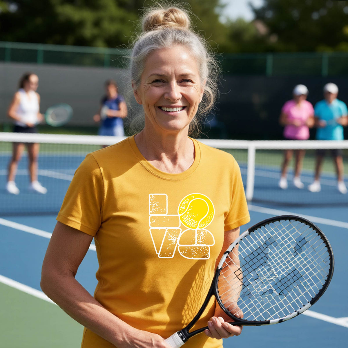 A woman wearing a gold short-sleeve t-shirt with a white and yellow tennis-themed graphic "LOVE" with a tennis ball replacing the O, with a tennis  court and players in the background.