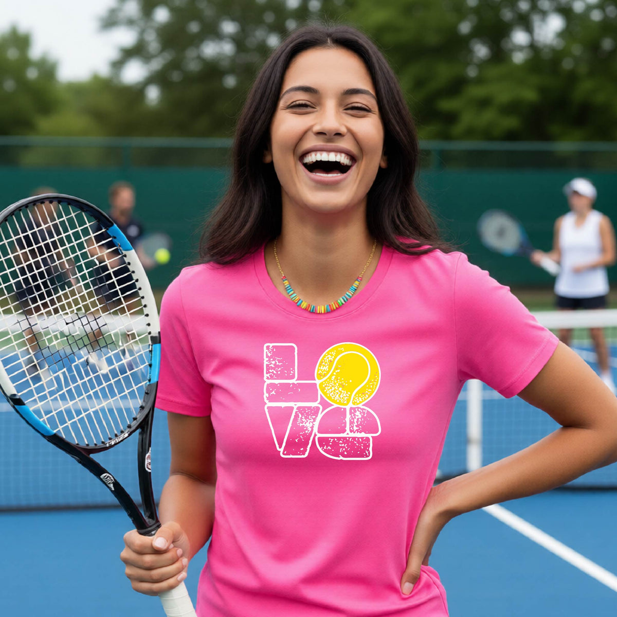 A woman wearing a pink short-sleeve t-shirt with a white and yellow tennis-themed graphic "LOVE" with a tennis ball replacing the O, with a tennis  court and players in the background.