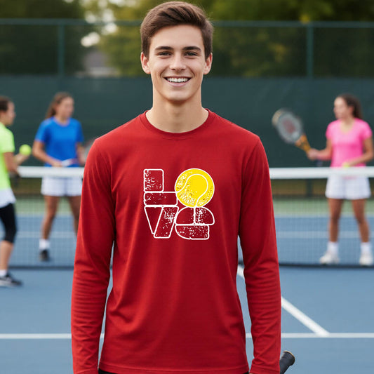 A smiling young man stands on a tennis court wearing a red long-sleeve shirt with a distressed white graphic spelling 'LOVE' and a yellow tennis ball motif. Blurred players are visible in the background on the court.