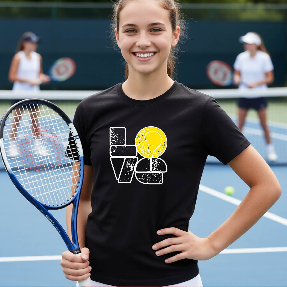 A smiling young tennis player stands on a blue court holding a tennis racket, wearing a black graphic T-shirt with a distressed white 'LOVE' design and a yellow tennis ball motif. Two blurred players are visible in the background on the court.