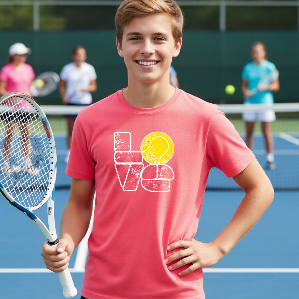 A smiling young tennis player stands on a blue court holding a tennis racket, wearing a coral colored graphic T-shirt with a distressed white 'LOVE' design and a yellow tennis ball motif. Two blurred players are visible in the background on the court.