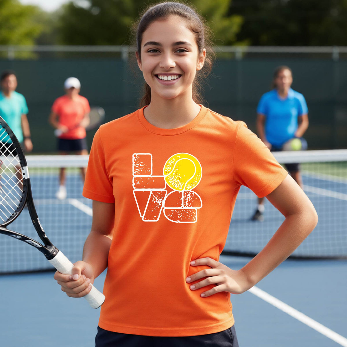 A smiling young tennis player stands on a blue court holding a tennis racket, wearing an orange graphic T-shirt with a distressed white 'LOVE' design and a yellow tennis ball motif. Two blurred players are visible in the background on the court.