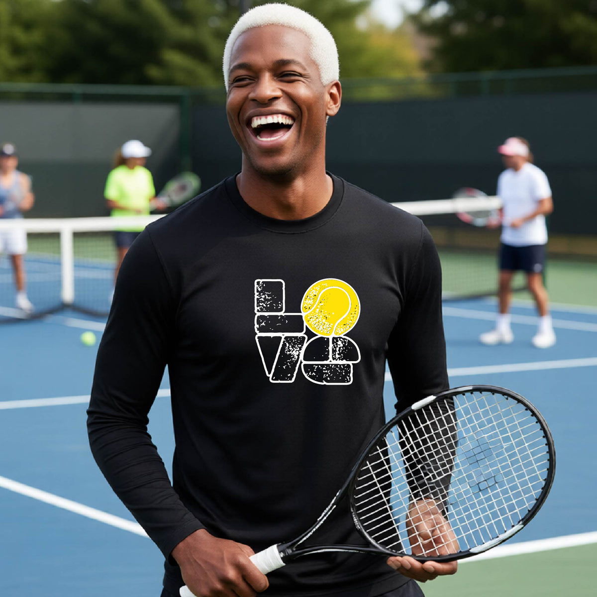 A man wearing a black long-sleeve t-shirt with a white and yellow tennis-themed graphic "LOVE" with a tennis ball replacing the O, with a tennis  court and players in the background.