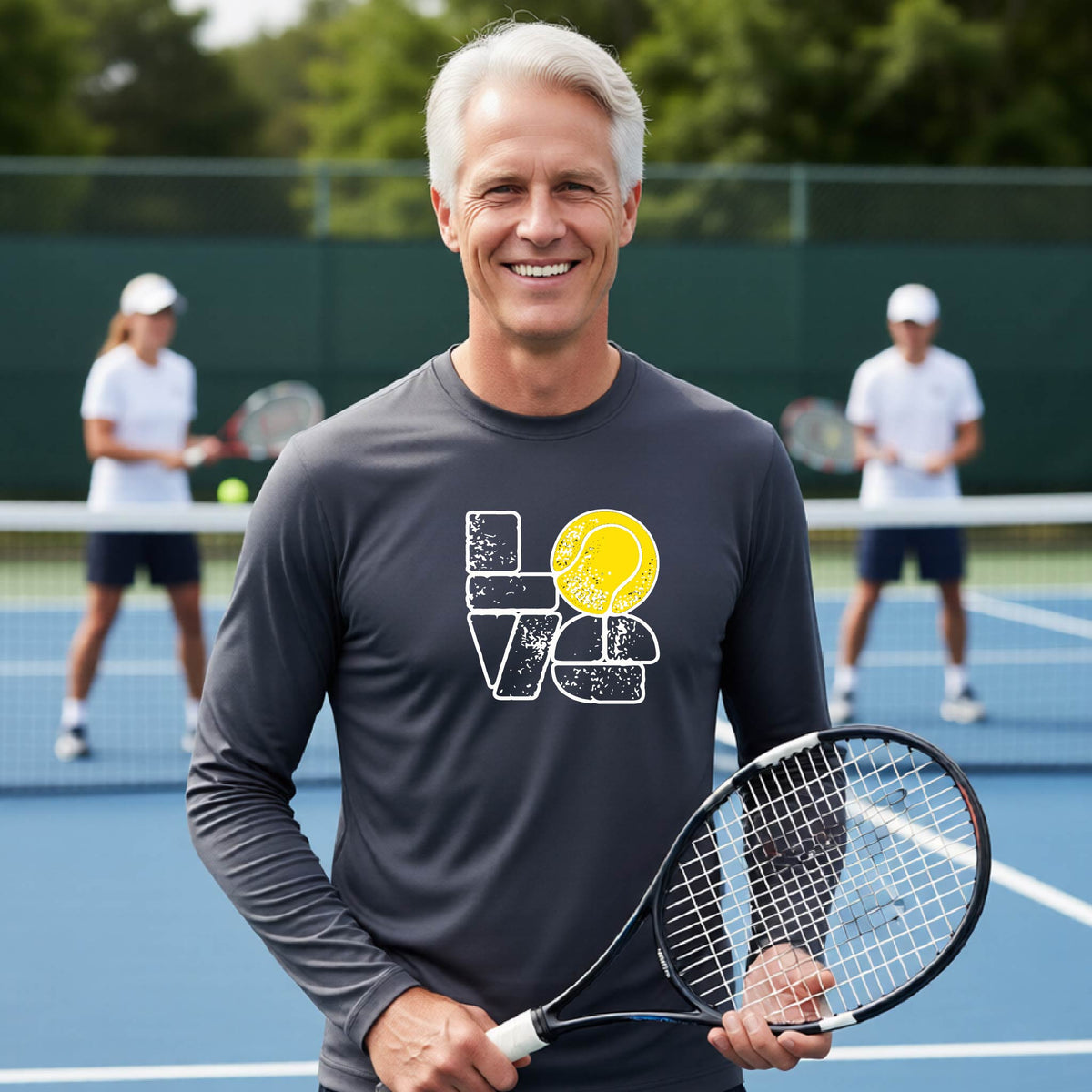A man wearing a dark gray long-sleeve t-shirt with a white and yellow tennis-themed graphic "LOVE" with a tennis ball replacing the O, with a tennis  court and players in the background.