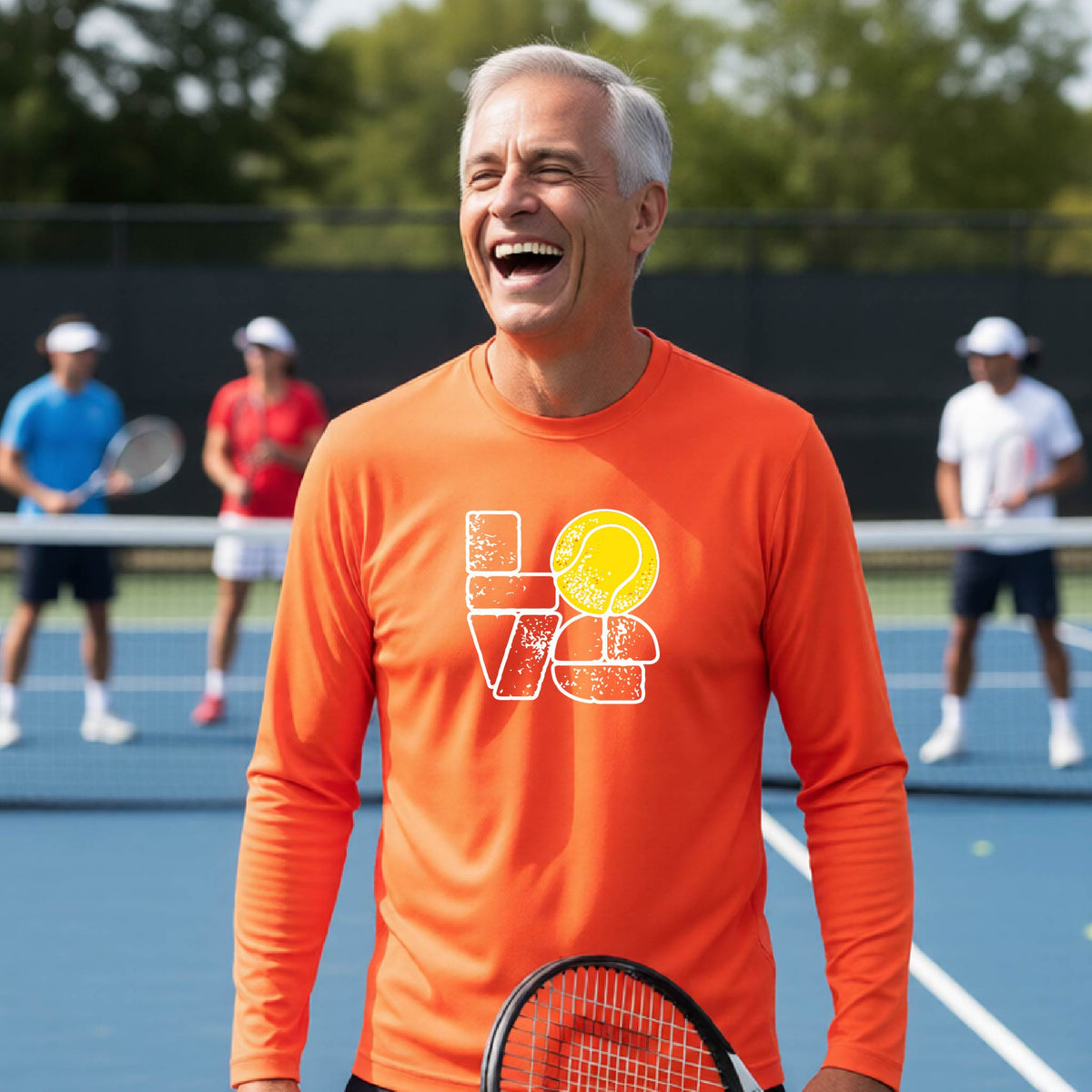 A man wearing an orange long-sleeve t-shirt with a white and yellow tennis-themed graphic "LOVE" with a tennis ball replacing the O, with a tennis  court and players in the background.