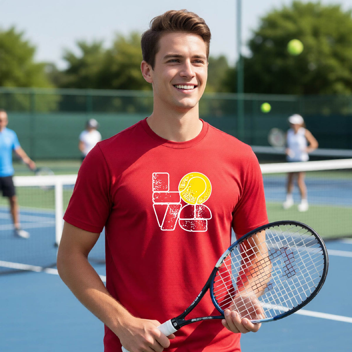 A man wearing a red short-sleeve t-shirt with a white and yellow tennis-themed graphic "LOVE" with a tennis ball replacing the O, with a tennis  court and players in the background.