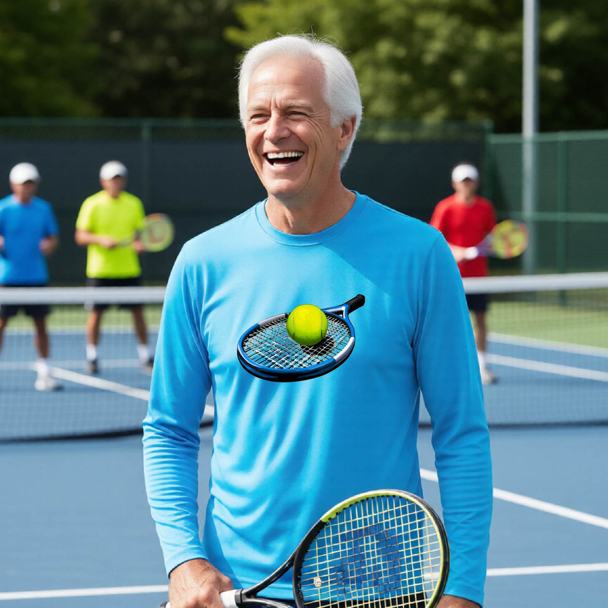A smiling older man on a tennis court wearing a bright blue long-sleeve shirt, holding a tennis racket, with tennis balls on the chest graphic area and other players blurred in the background.