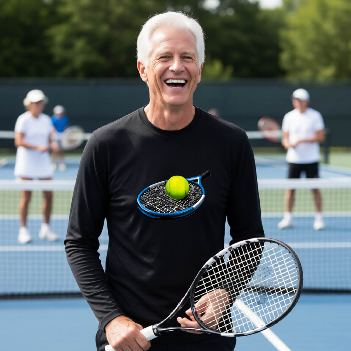 A smiling older man on a tennis court wearing a black long-sleeve shirt with a graphic of a tennis racket and yellow tennis ball on the chest, holding a tennis racket. Other players are blurred in the background.