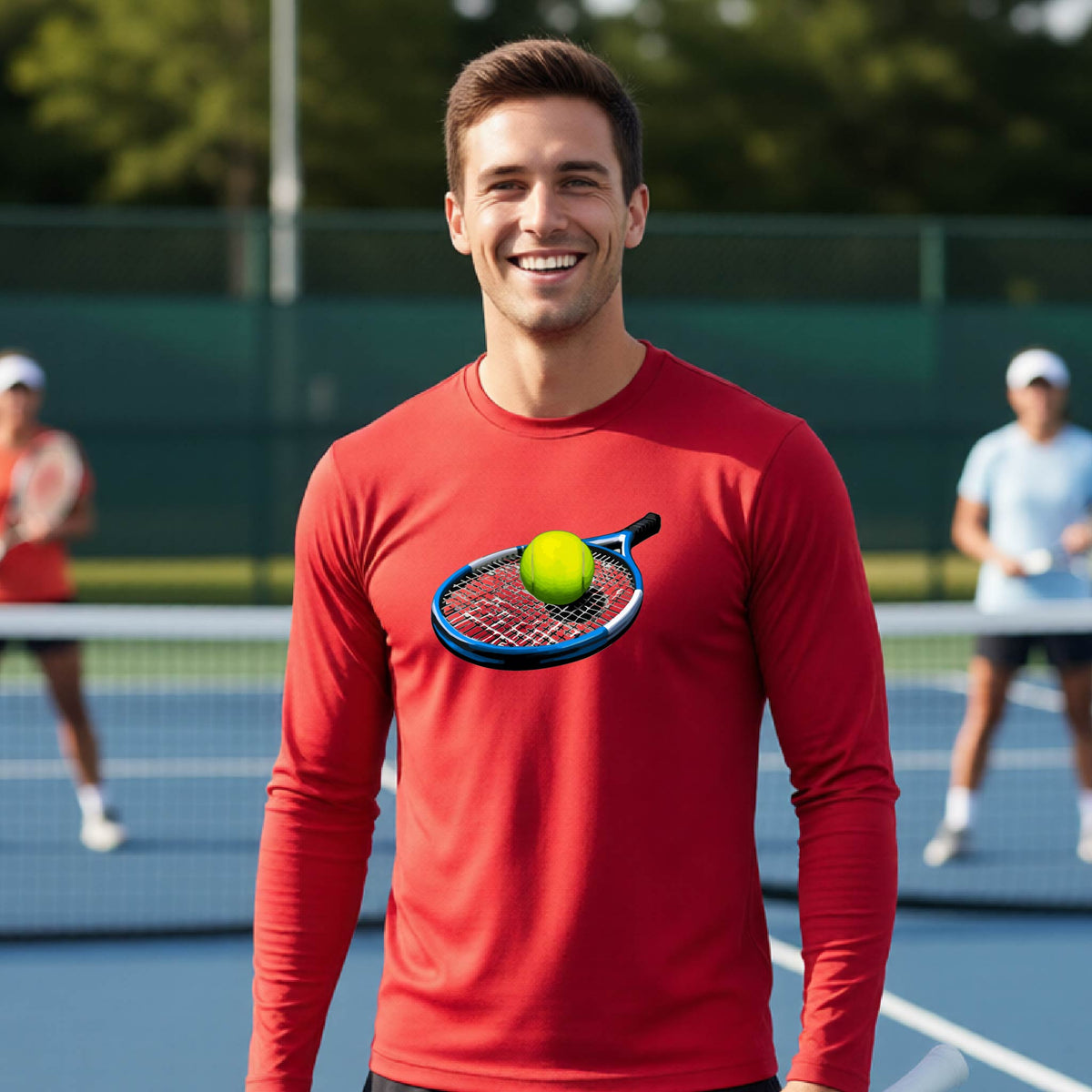 A smiling man on a tennis court wearing a red long-sleeve shirt with a graphic of a tennis racket and yellow tennis ball on the chest, with blurred players in the background.