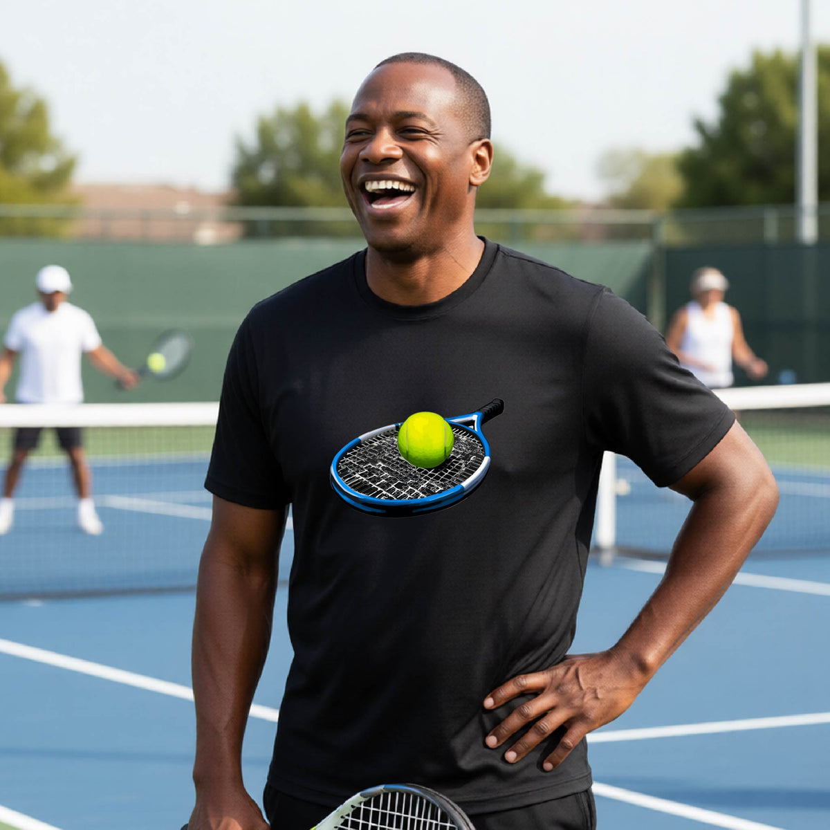 A man laughing on a tennis court wearing a black t-shirt with a graphic of a tennis racket and yellow tennis ball on the chest.