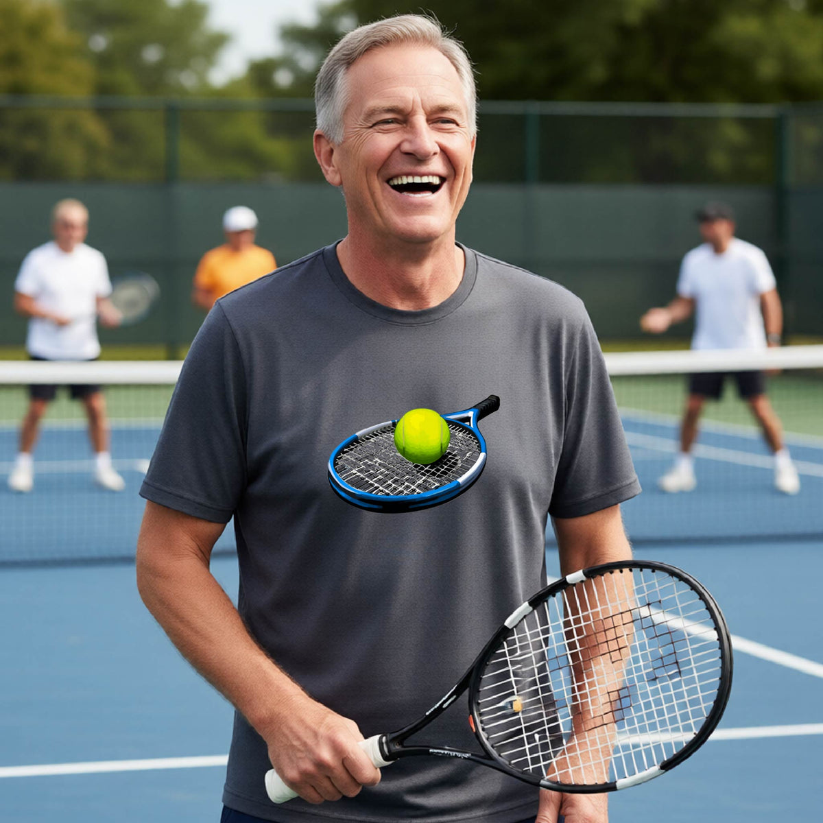 A smiling older man on a tennis court wearing a dark gray t-shirt with a graphic of a tennis racket and tennis ball, holding a tennis racket. Two other players are blurred in the background on the court.