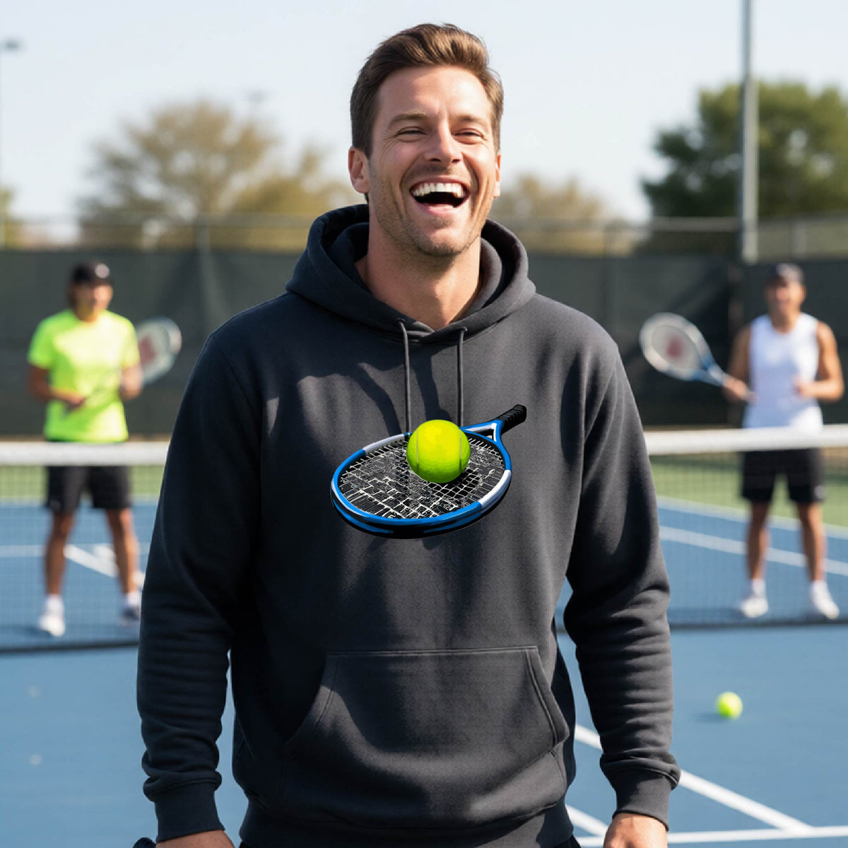 A man laughing on a tennis court wearing a dark gray hoodie with a graphic of a tennis racket and tennis ball on the chest, with other players blurred in the background.