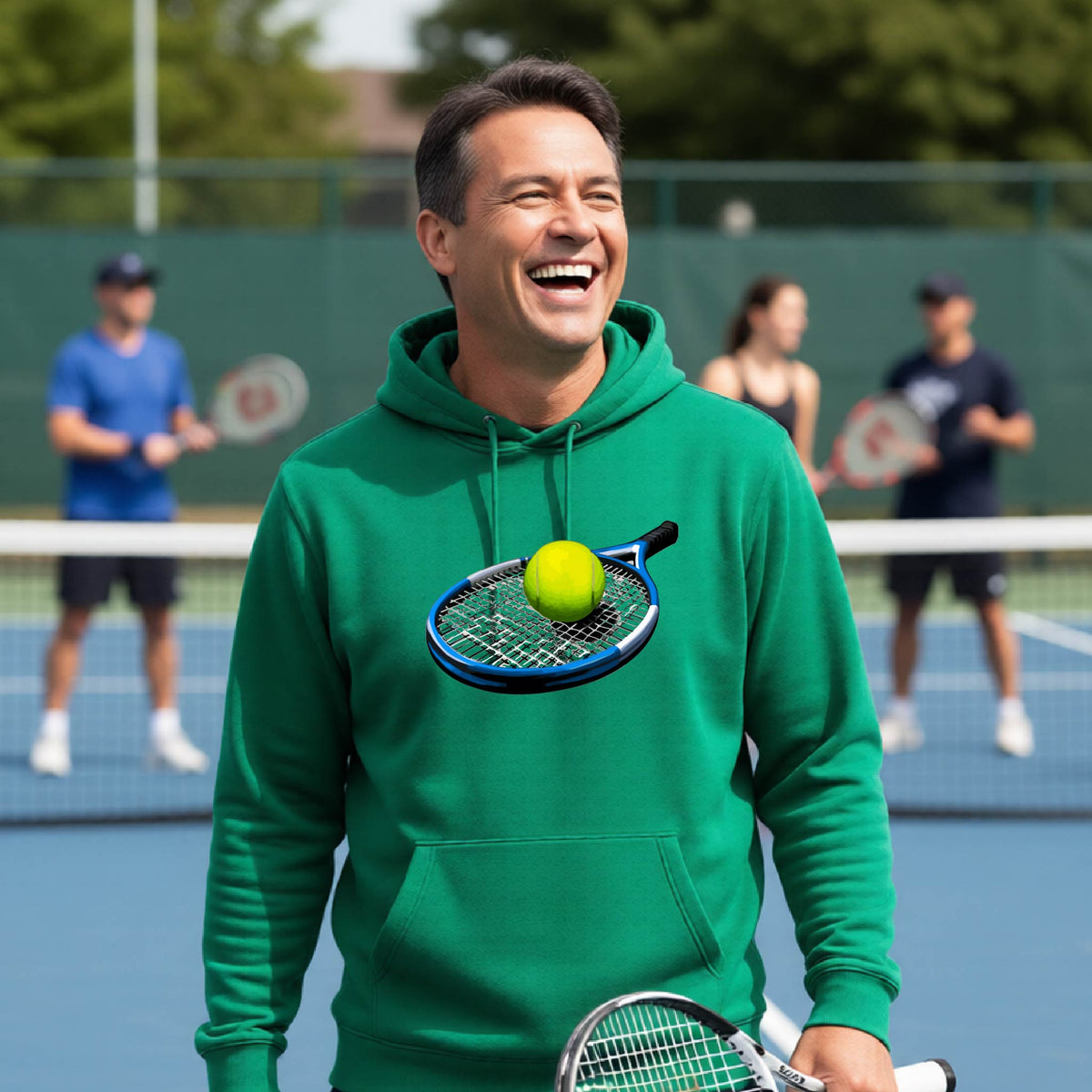 A man on a tennis court wearing a bright green hoodie with a graphic of a tennis racket and tennis ball on the chest, holding a tennis racket and smiling broadly. Other players are blurred in the background.