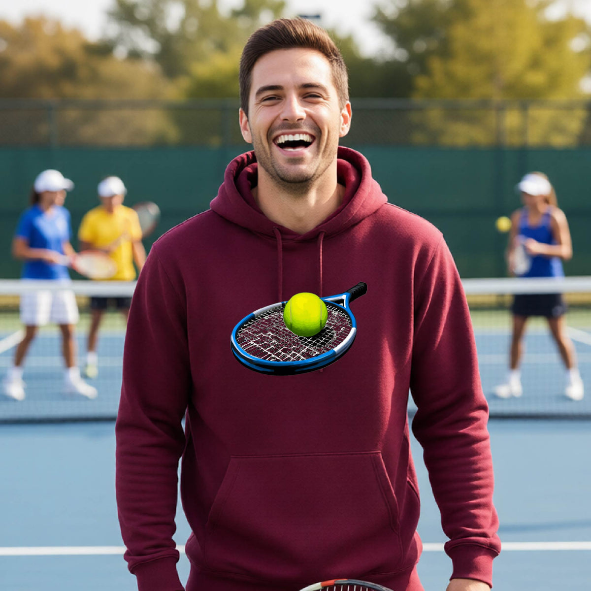 A man wearing a maroon hoodie with a graphic of a tennis racket and yellow tennis ball stands on a tennis court, with other players blurred in the background.