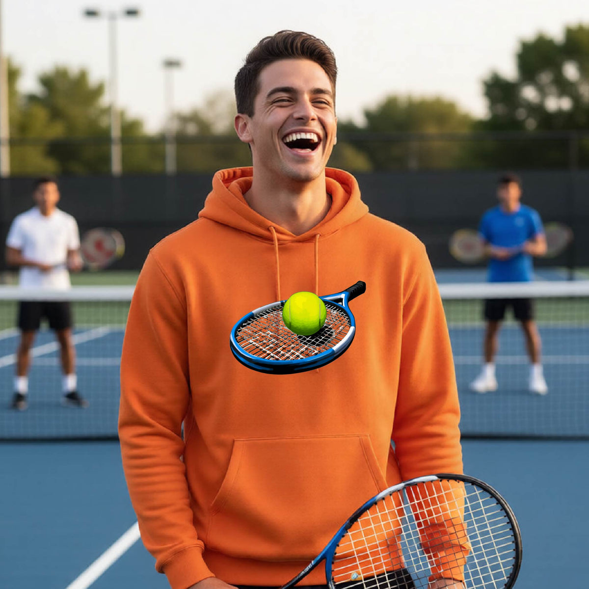 A man laughing on a tennis court wearing an orange hoodie with a graphic of a tennis racket and yellow tennis ball on the chest, holding a tennis racket.