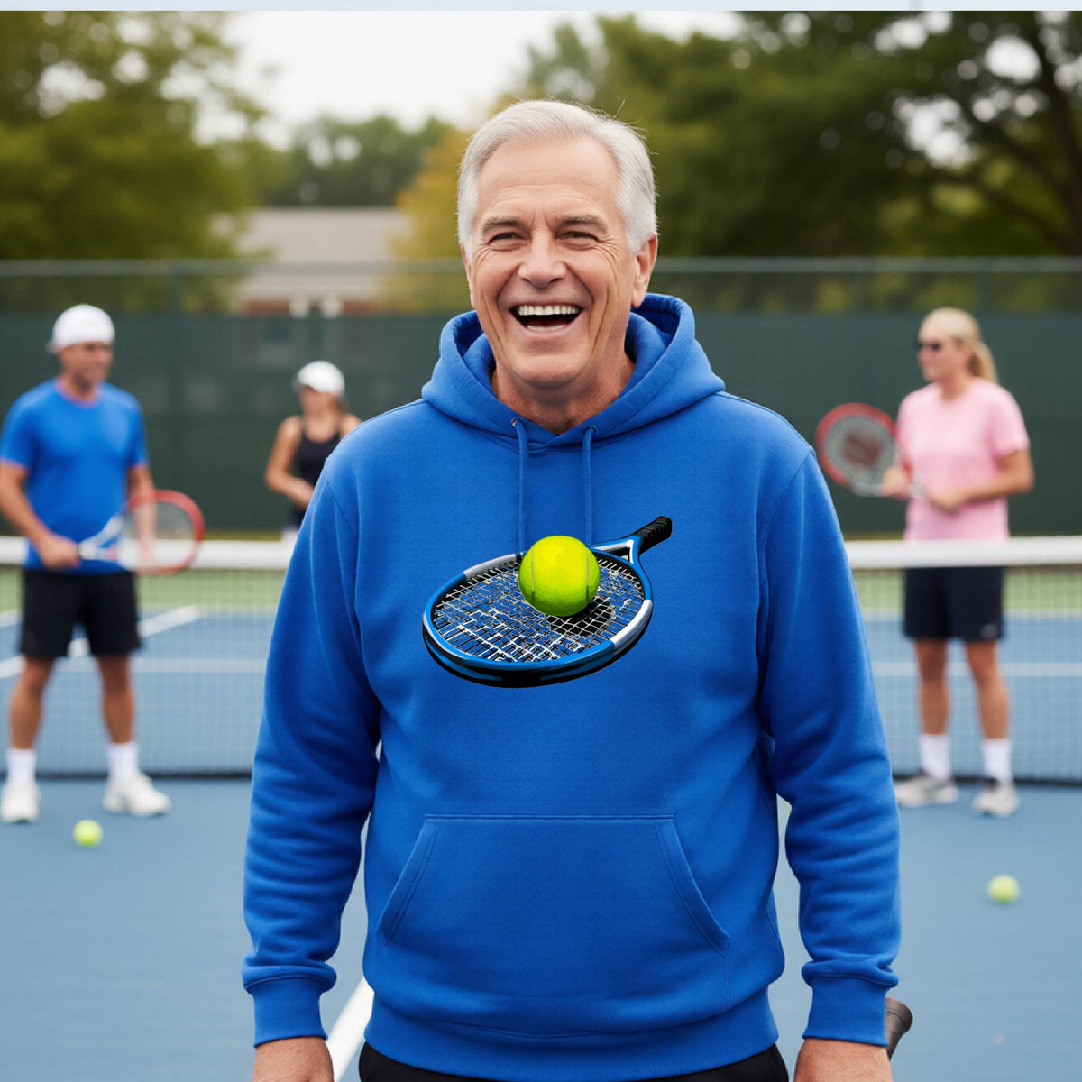 A smiling older man wearing a bright blue hoodie stands on a tennis court, with a tennis racket and tennis ball printed on the chest. Other tennis players are blurred in the background.