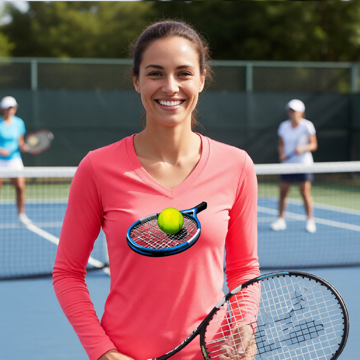 A smiling woman on a tennis court wearing a coral pink long-sleeve V-neck athletic shirt with a graphic of a tennis racket and tennis ball on the chest, holding a tennis racket. Two other players are blurred in the background.