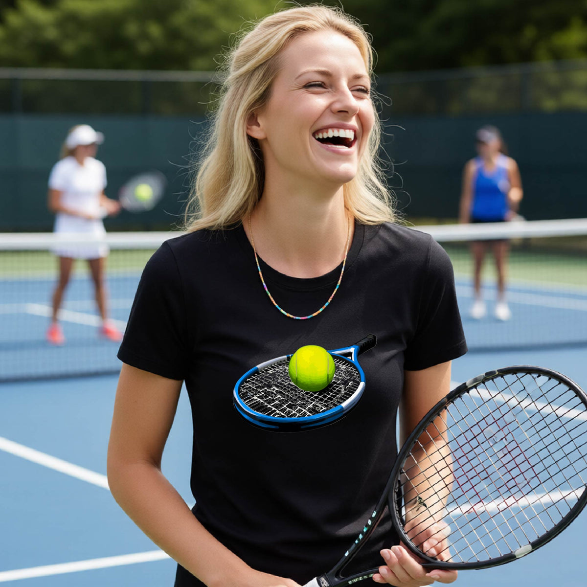 A woman laughing on a tennis court wearing a black t-shirt with a graphic of a tennis racket and yellow tennis ball. She is holding a tennis racket, with other players and the court visible in the background.