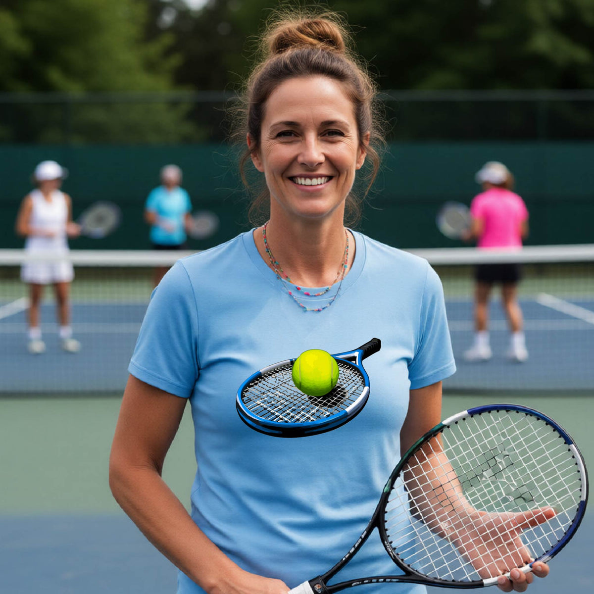 A smiling woman on a tennis court wearing a light blue t-shirt with a graphic of a tennis racket and tennis ball on the chest, holding a tennis racket. Other players are blurred in the background.