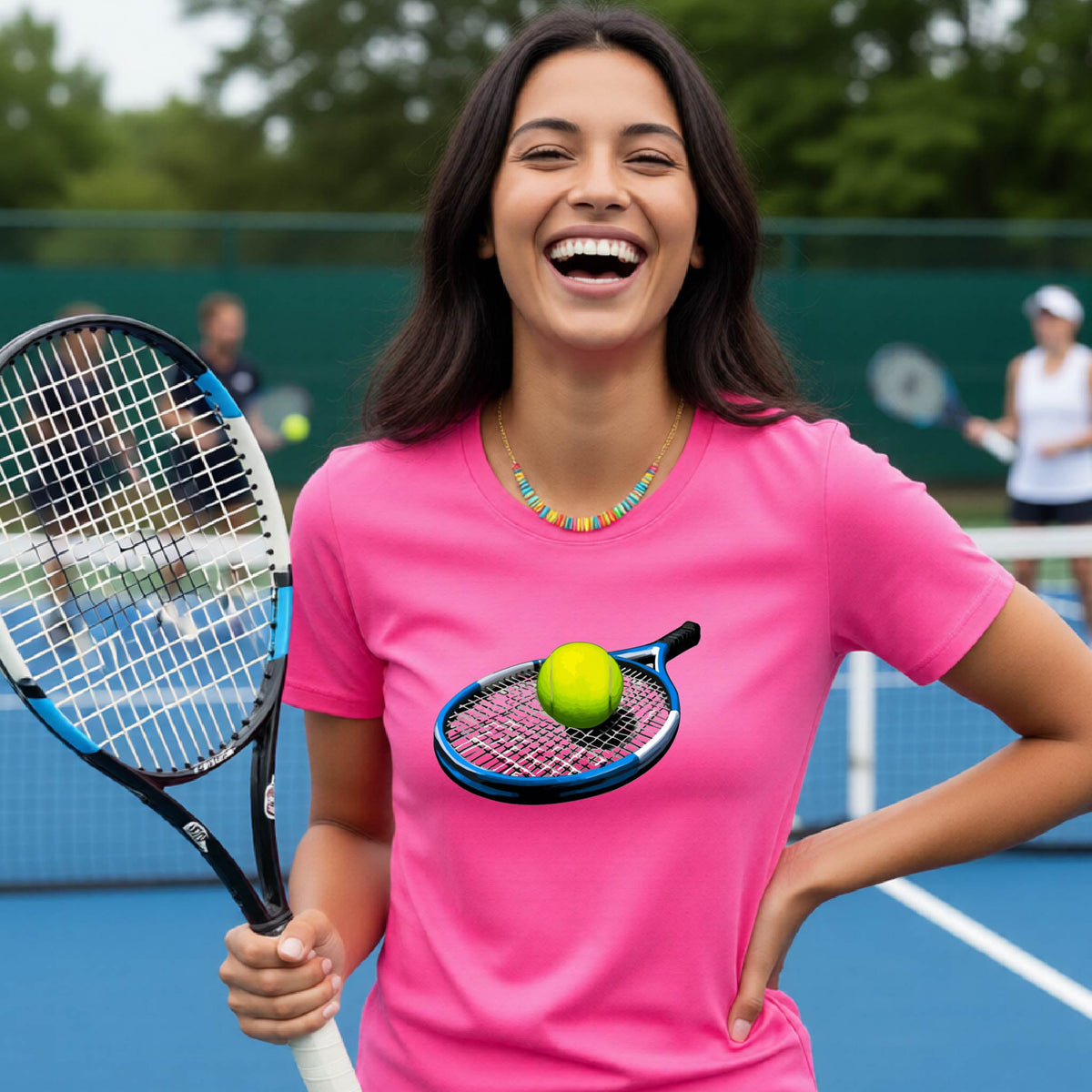 A woman laughing on a tennis court wearing a bright pink t-shirt with a graphic of a tennis racket and tennis ball on the chest, holding a tennis racket.