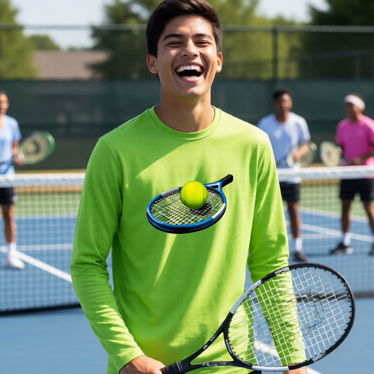 A smiling young man stands on a tennis court wearing a lime green long-sleeve shirt with a graphic of a tennis racket and yellow tennis ball on the front. She holds a tennis racket, with other players blurred in the background.