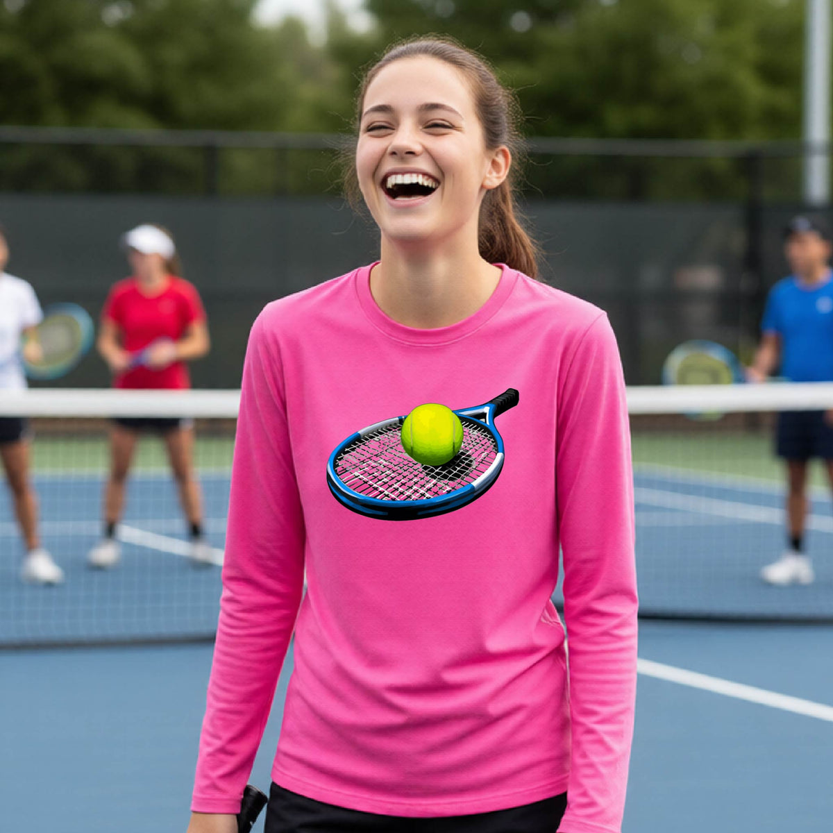 A smiling young woman stands on a tennis court wearing a bright pink long-sleeve shirt with a graphic of a tennis racket and yellow tennis ball on the front. She holds a tennis racket, with other players blurred in the background.