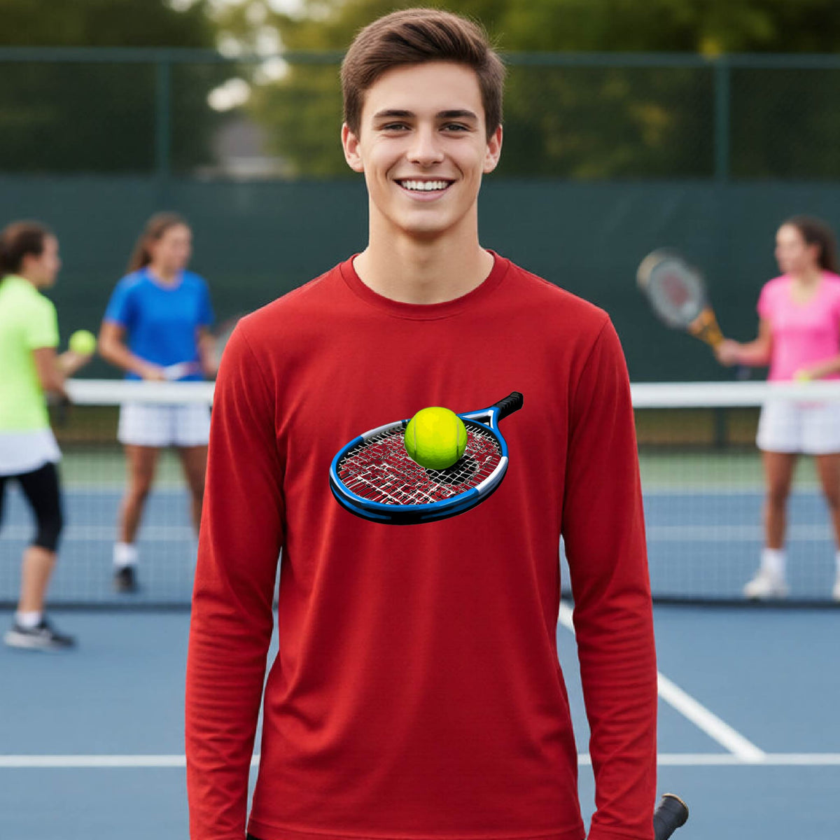A smiling young man stands on a tennis court wearing a red long-sleeve shirt with a graphic of a tennis racket and yellow tennis ball on the chest. Other players are blurred in the background.