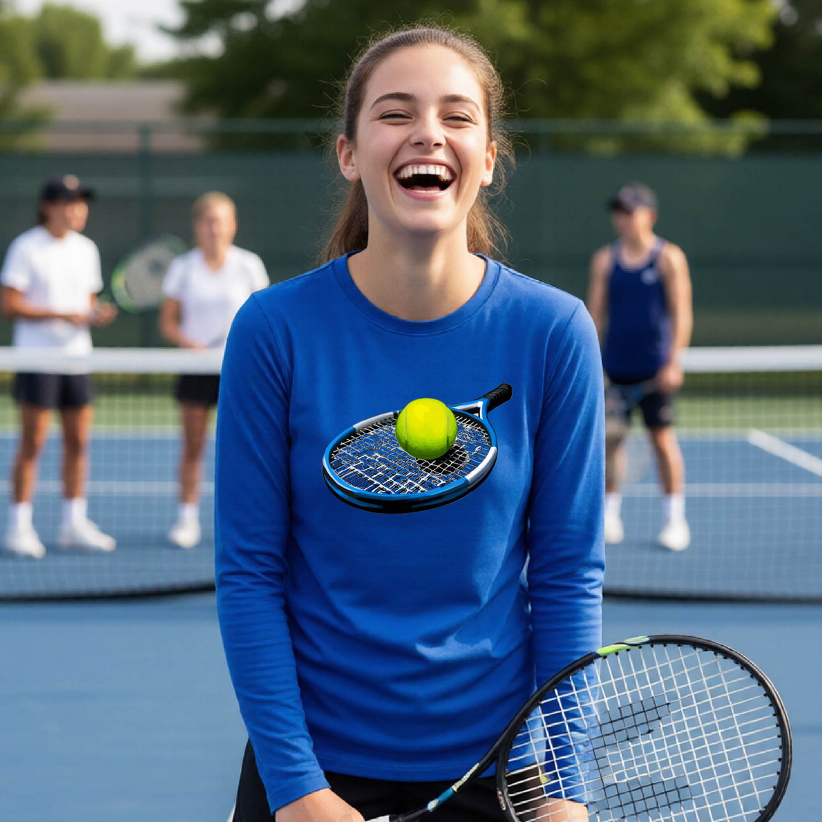 A young woman laughing on an outdoor tennis court, wearing a bright blue long-sleeve shirt with a graphic of a tennis racket and yellow tennis ball on the chest, holding a tennis racket.
