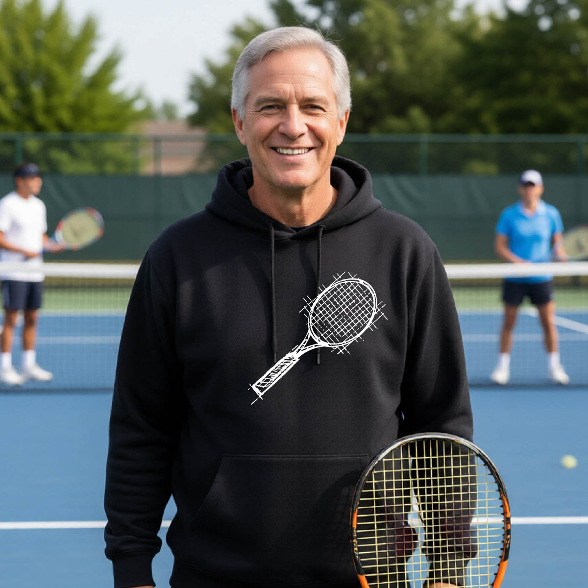 A man wearing a black hoodie with a white tennis racket retro graphic on the chest, standing on a blue tennis court with players blurred in the background.