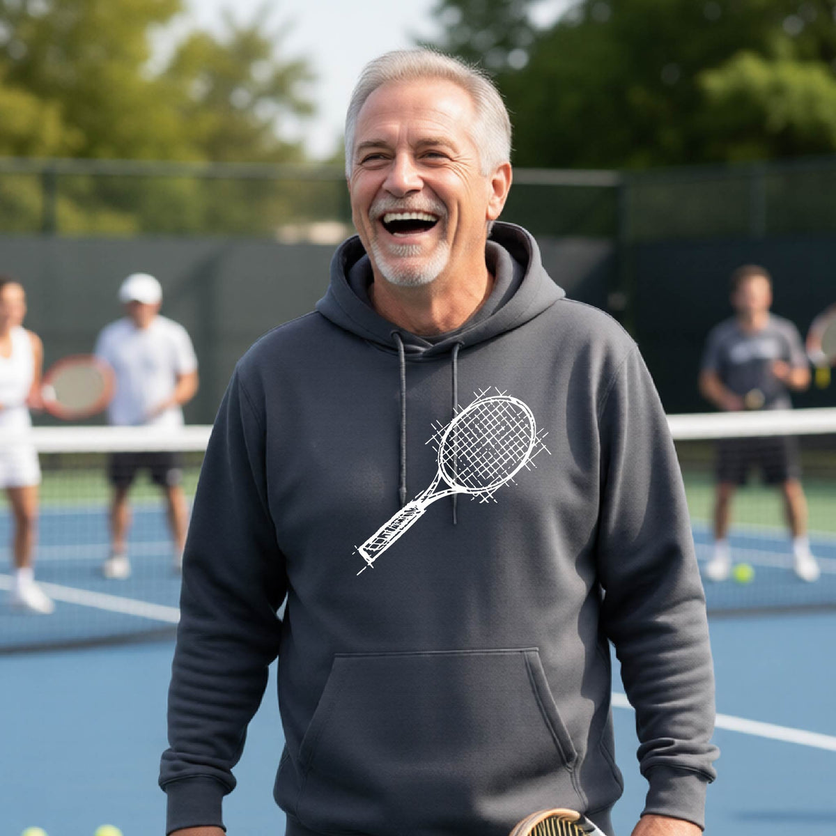 A man wearing a gray hoodie with a white tennis racket retro graphic on the chest, standing on a blue tennis court with players blurred in the background.