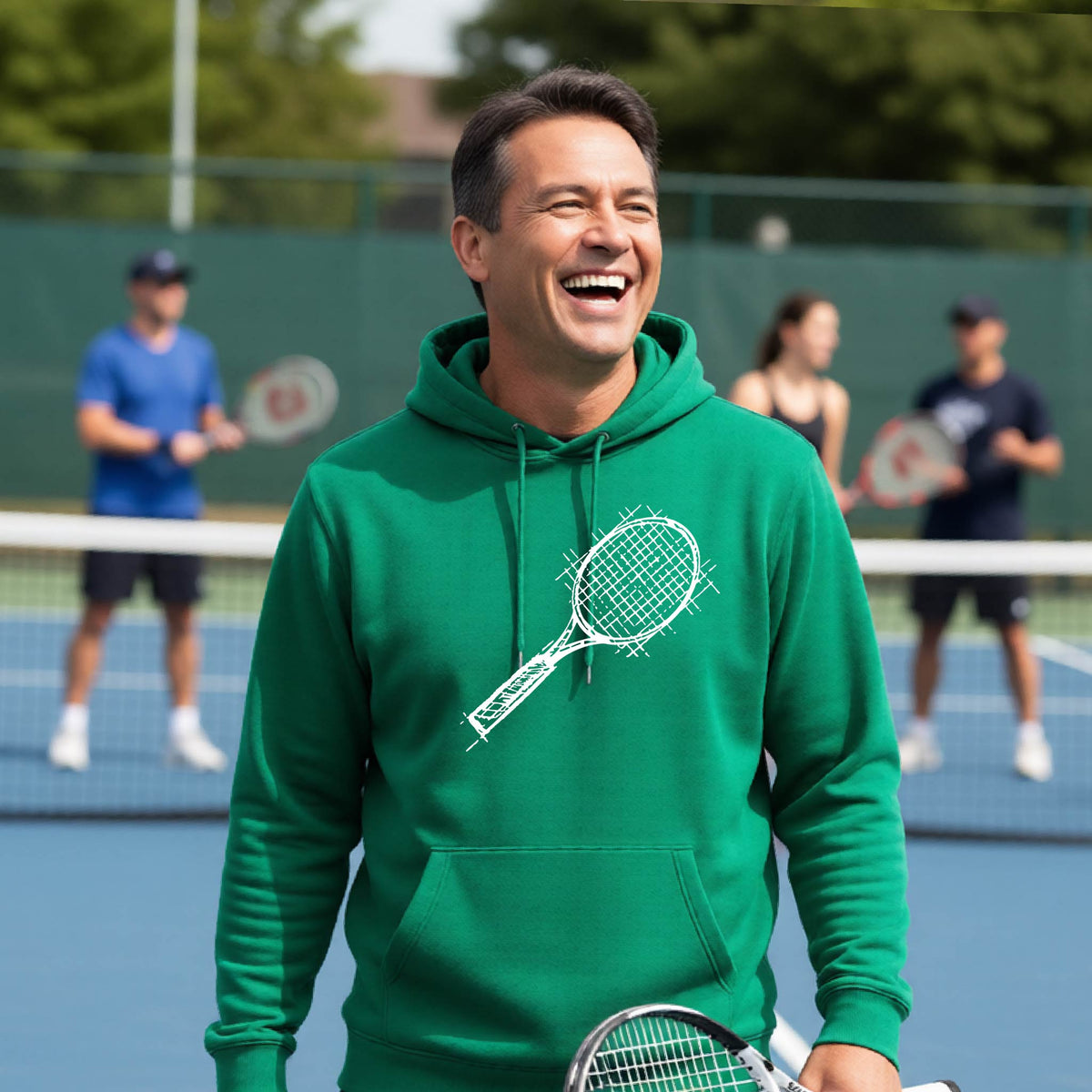 A man wearing a green hoodie with a white tennis racket retro graphic on the chest, standing on a blue tennis court with players blurred in the background.