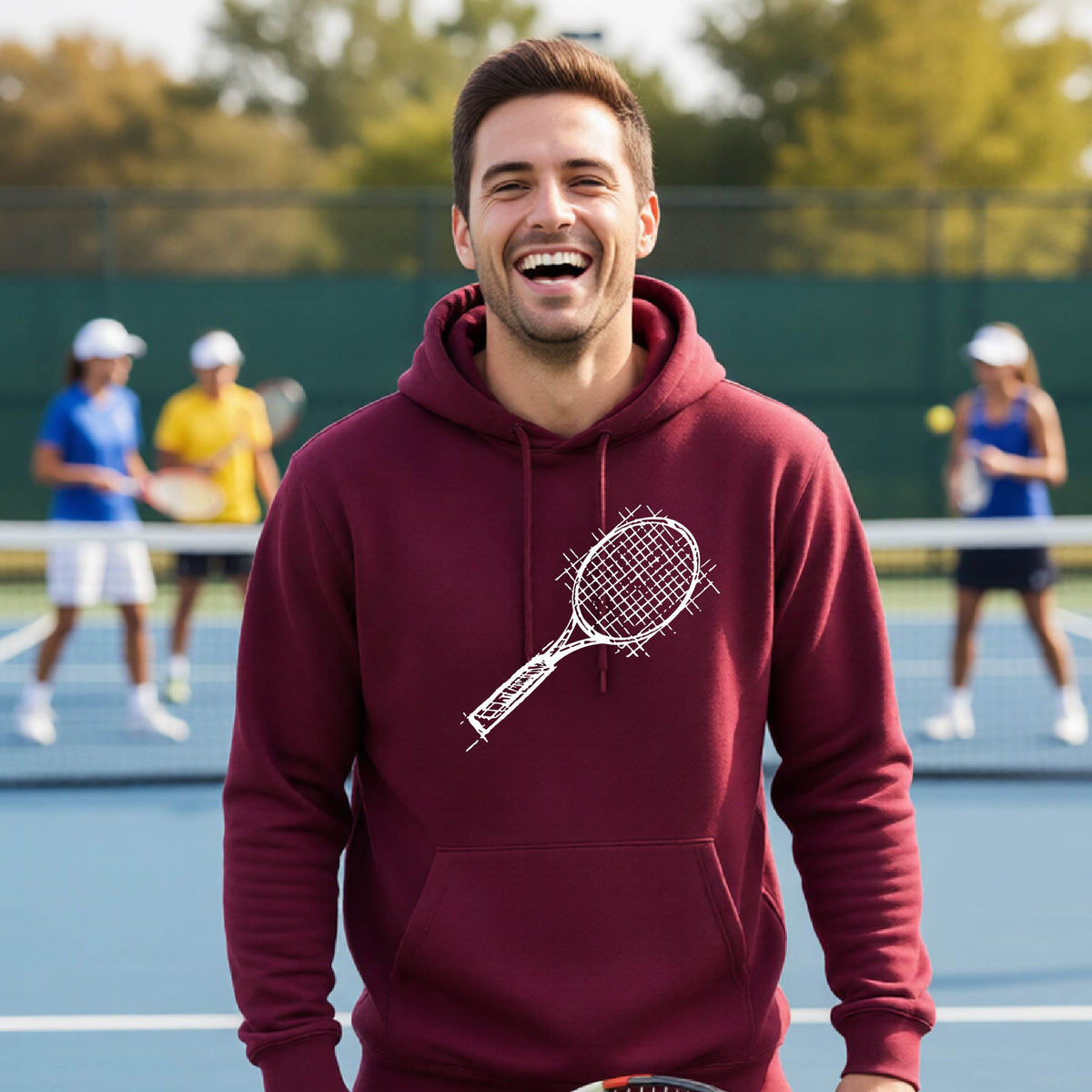 A man wearing a maroon hoodie with a white tennis racket retro graphic on the chest, standing on a blue tennis court with players blurred in the background.