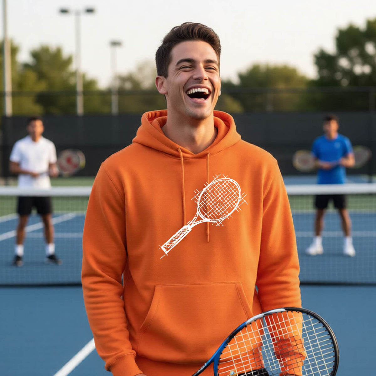 A man wearing an orange hoodie with a white tennis racket retro graphic on the chest, standing on a blue tennis court with players blurred in the background.