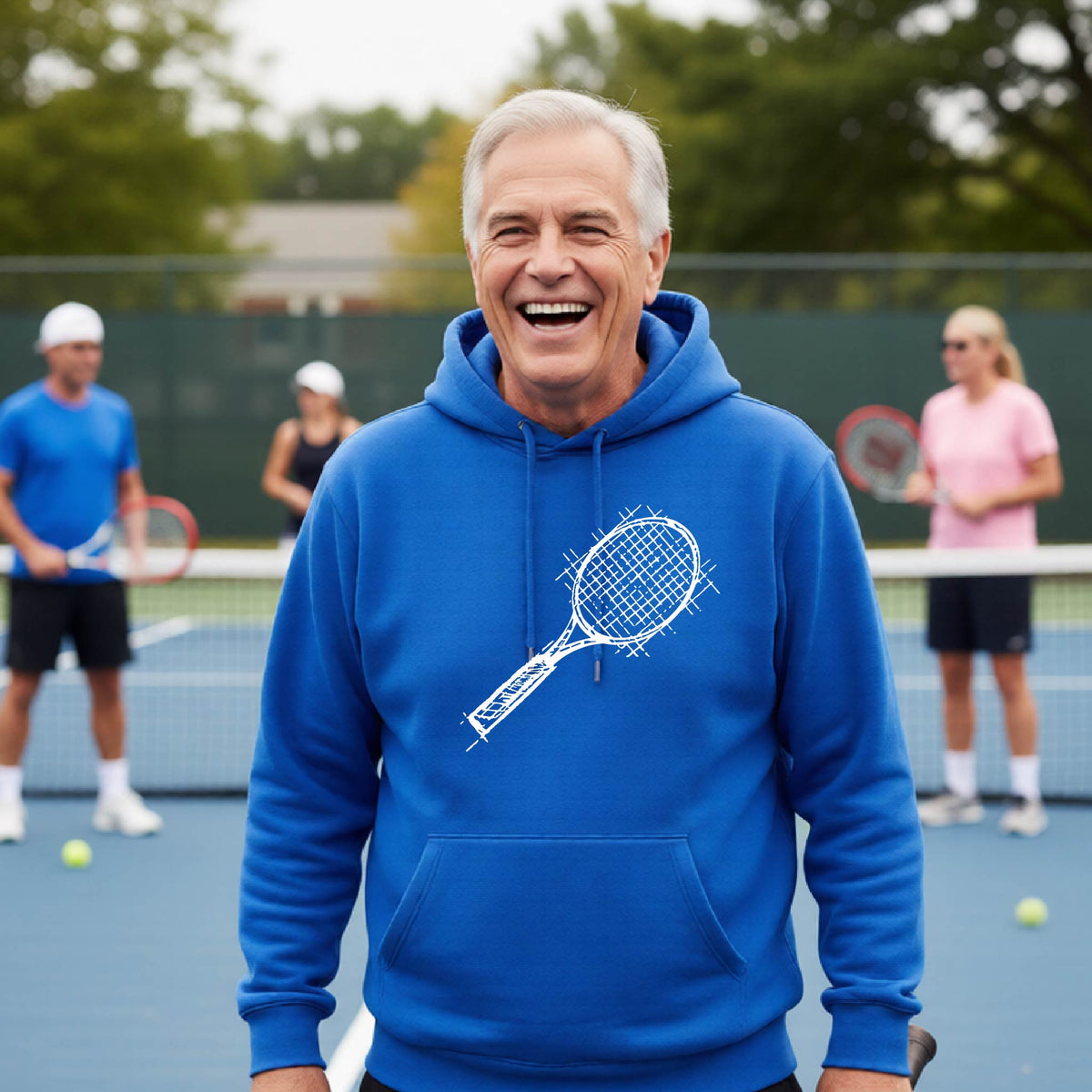 A man wearing a royal blue hoodie with a white tennis racket retro graphic on the chest, standing on a blue tennis court with players blurred in the background.