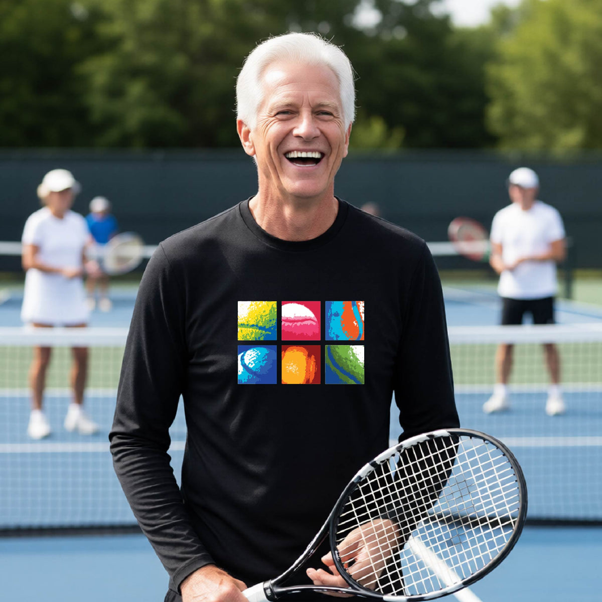 A smiling man on a blue tennis court holds a tennis racket and wears a black long-sleeve T-shirt with a colorful 2x3 grid graphic on the chest of tennis balls. Other tennis players are blurred in the background.