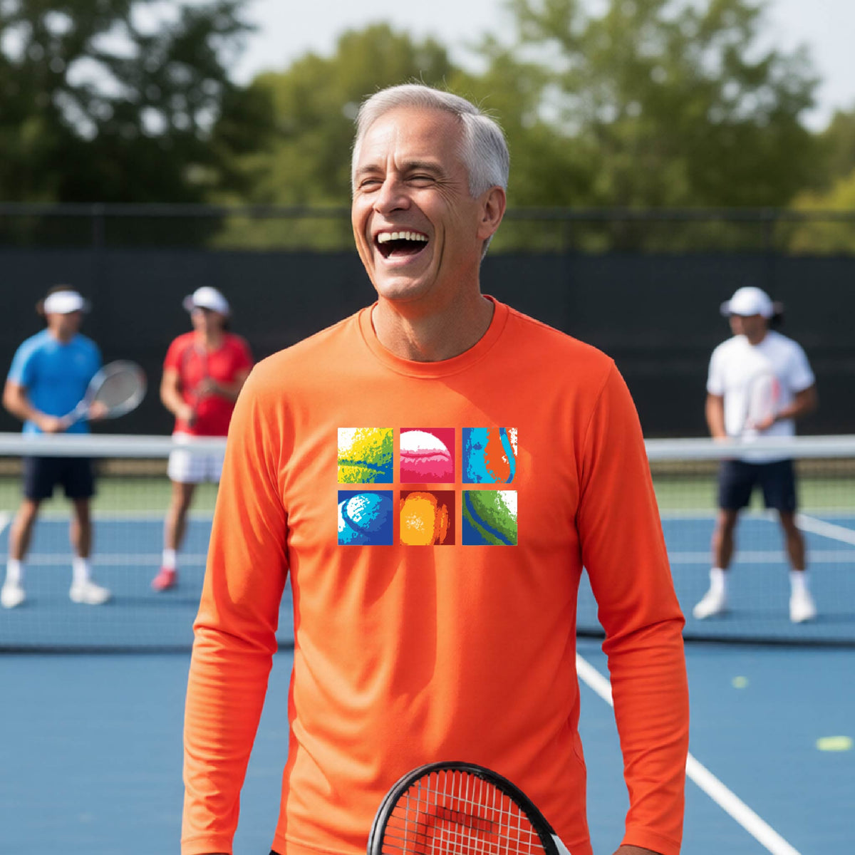 A smiling man on a blue tennis court holds a tennis racket and wears an orange long-sleeve T-shirt with a colorful 2x3 grid graphic on the chest of tennis balls. Other tennis players are blurred in the background.