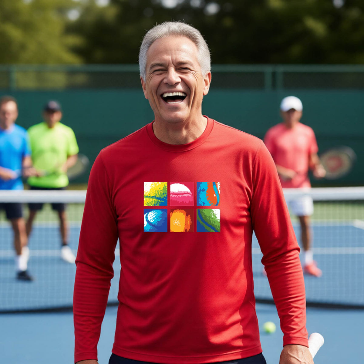 A smiling man on a blue tennis court holds a tennis racket and wears a red long-sleeve T-shirt with a colorful 2x3 grid graphic on the chest of tennis balls. Other tennis players are blurred in the background.