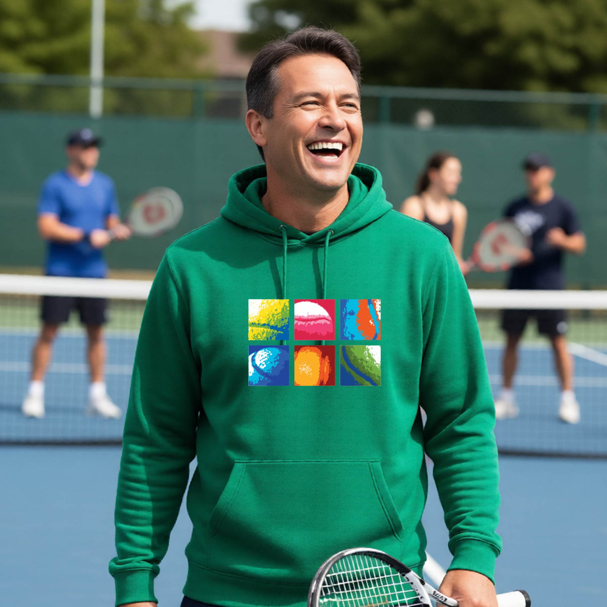 A man wearing a green hoodie with a colorful 3x3 abstract graphic print on tennis balls on the chest, standing on a blue tennis court with other players blurred in the background.