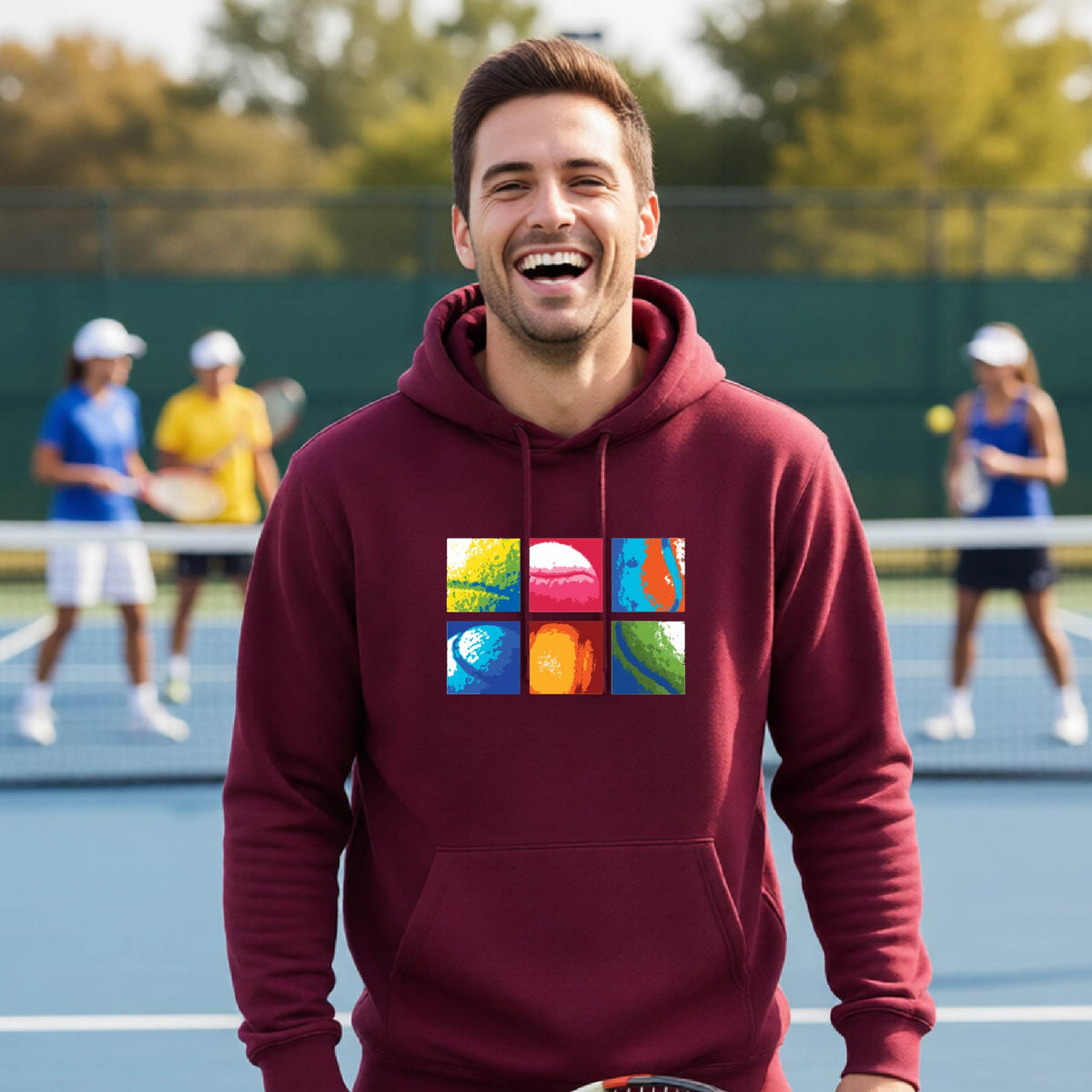 A man wearing a maroon hoodie with a colorful 3x3 abstract graphic print on tennis balls on the chest, standing on a blue tennis court with other players blurred in the background.