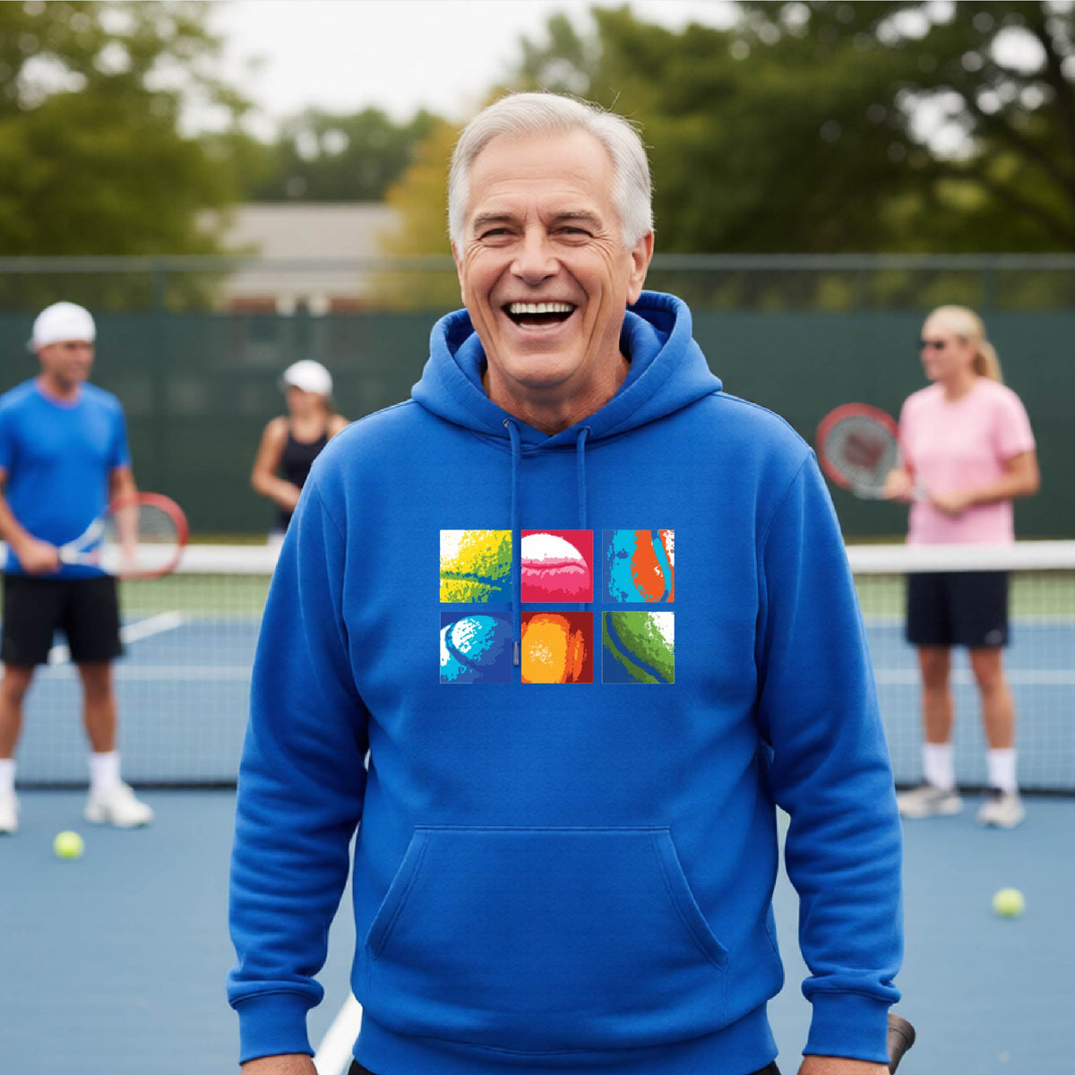 A man wearing a royal blue hoodie with a colorful 3x3 abstract graphic print on tennis balls on the chest, standing on a blue tennis court with other players blurred in the background.