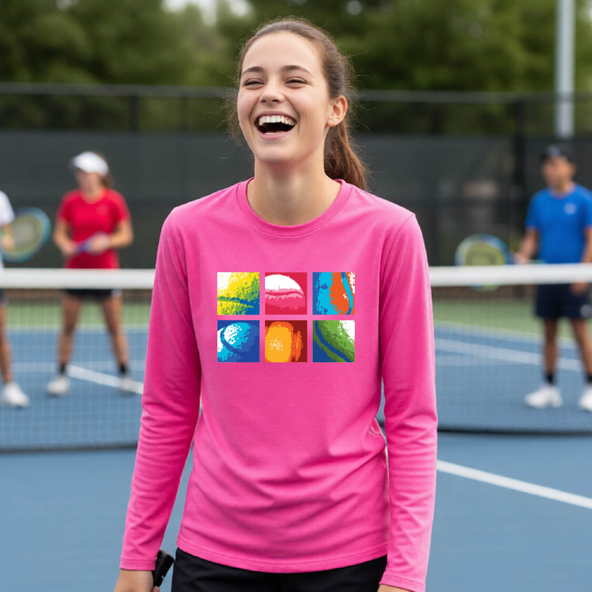 A woman laughing on a tennis court wearing a bright pink long-sleeve shirt with a colorful graphic print on the chest.
