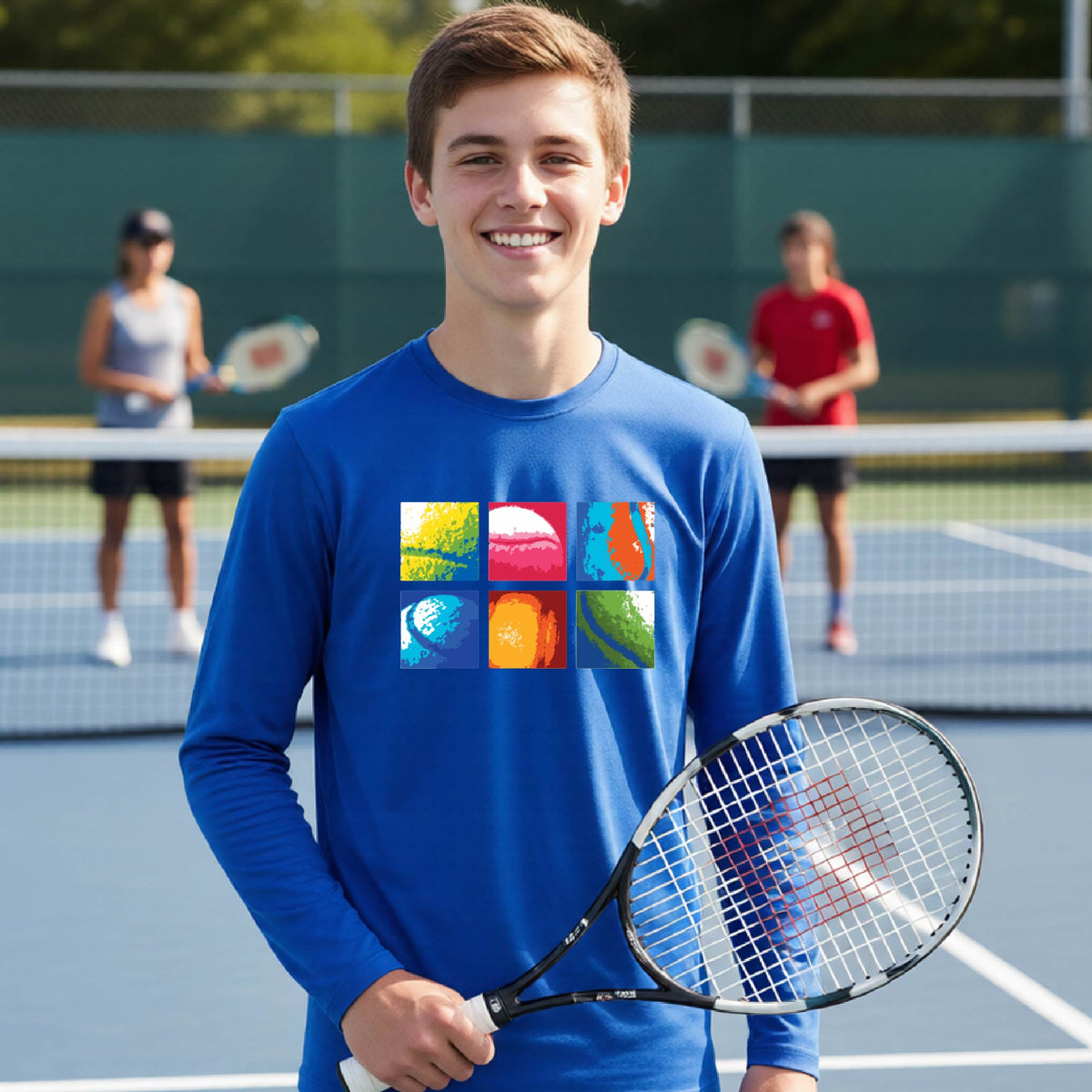 A smiling young man on a tennis court wearing a royal blue long-sleeve shirt with a colorful graphic of six tennis balls in different colors. Two other players are visible in the background on the court.