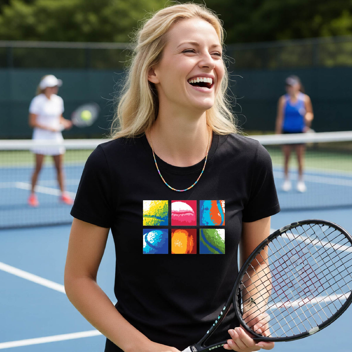 A smiling woman on a blue tennis court holds a tennis racket and wears a black short-sleeve T-shirt with a colorful 2x3 grid graphic on the chest of tennis balls. Other tennis players are blurred in the background.